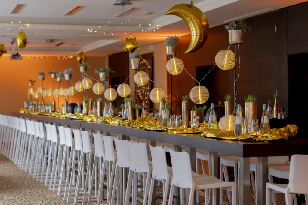 Foto bei Tageslicht aufgenommen von den Veranstaltungsräumen D1, D2 und D3 zu sehen die Partybestuhlung für die anstehende Tastetival Wein und Genussparty am Abend. Hochtische und Hochstühle in den Räumen zu einer langen Tafel zusammengestellt. Von der Decke hängen Ballons in Formen von Mond und Sternen, Die lange Tafel ist golden Dekoriert mit einer Ballonartigen Lichterkette an den hohen Gusseisernen Dekoständern befestigt