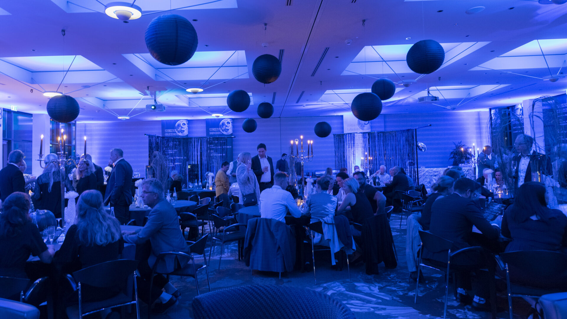 Aufnahme am Abend der Veranstaltungsräume E1, E2 und E3 kombiniert an unserer großen Wein und Genussparty. Von der Decke hängen PomPoms im Vordergrund zu sehen viele Gäste die an runden Bankett Tischen sitzten