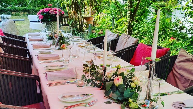 Eingedeckte lange Tafel in unserem Atrium Garten mit viel Grün und rosa Pflanzen im Hintergrund