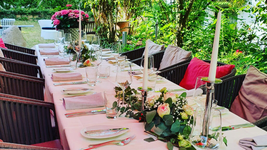 Eingedeckte lange Tafel in unserem Atrium Garten mit viel Grün und rosa Pflanzen im Hintergrund