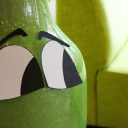 Foto von Blumenvase im Hintergrund leicht verschwommen ein grüner Sessel. Auf der Vase aufgeklebt sind zwei Augen die nach links schauen.