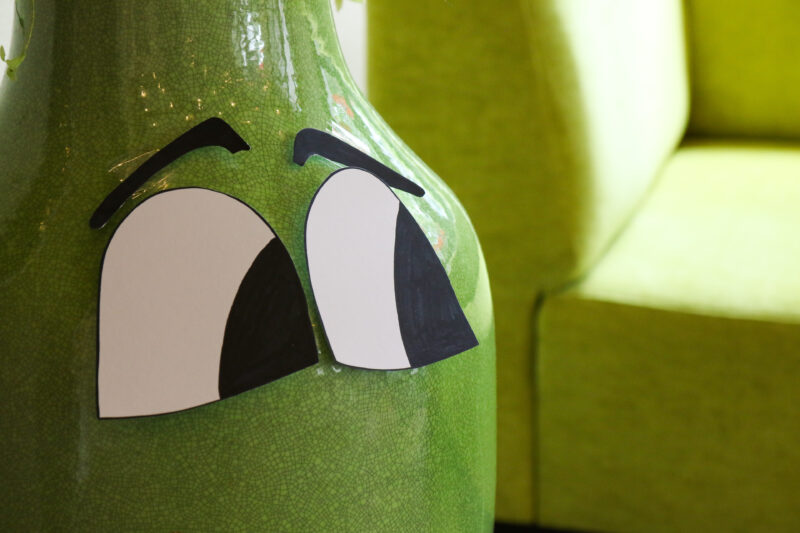 Foto von Blumenvase im Hintergrund leicht verschwommen ein grüner Sessel. Auf der Vase aufgeklebt sind zwei Augen die nach links schauen.