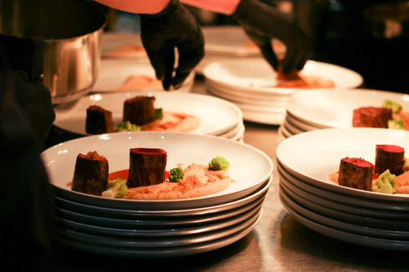 Detailfoto beim Anrichtes eines Hauptganges innerhalb eines festlichen Menüs. Auf dem Teller Selleriecreme, Brokolie und Ochsenbacken die von einem Koch mit Salz bestreut werden