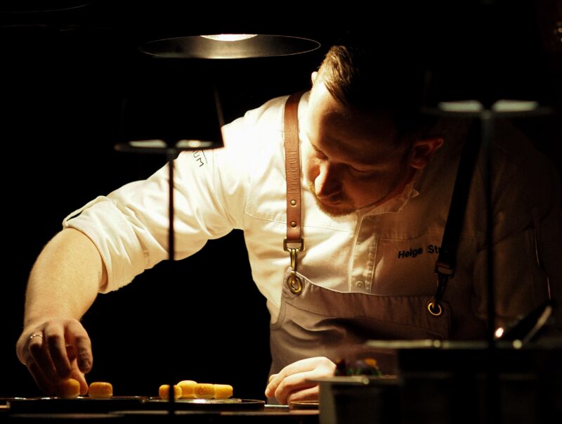 Koch beim Anrichten eines Desserts, er schaut konzentriert, am Bildrand sind Wärmelampen