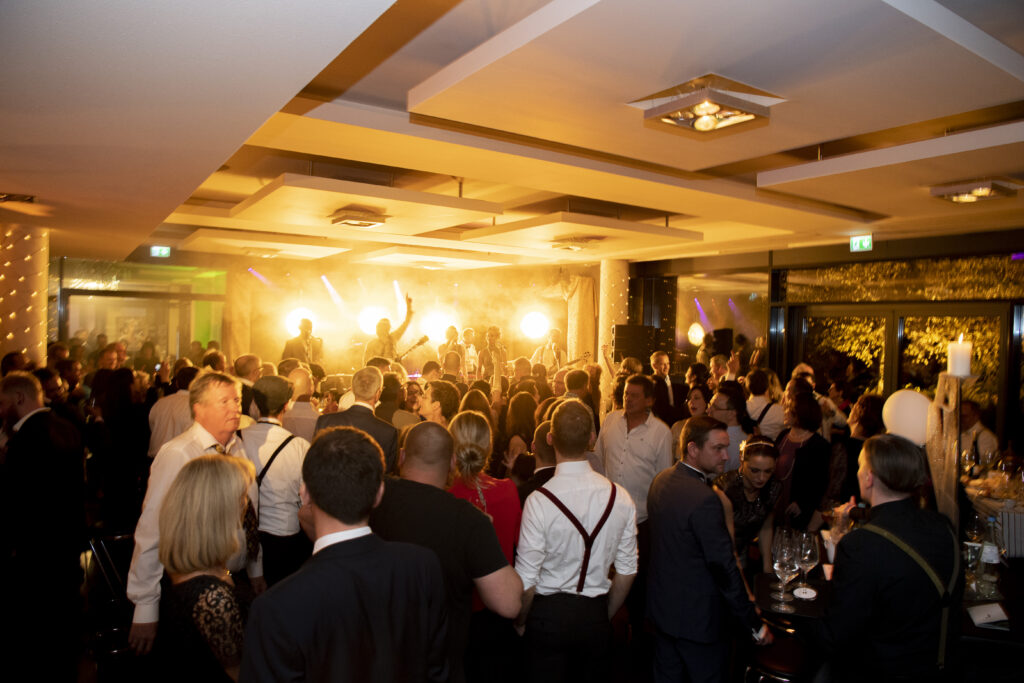Partybild aufgenommen aufunserer großen Tastetival Wein und Genussparty im Foyer vor den Veranstaltungsräumen D1-D3 im Hintergrund eine Band auf der Bühne zu sehen im Vordergrund eine Menschenmenge beim Tanzen