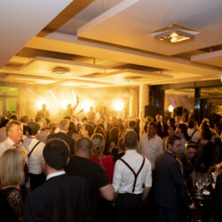 Partybild aufgenommen aufunserer großen Tastetival Wein und Genussparty im Foyer vor den Veranstaltungsräumen D1-D3 im Hintergrund eine Band auf der Bühne zu sehen im Vordergrund eine Menschenmenge beim Tanzen